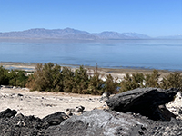 Zoom view overlooking a black outcrop with the shoreline of the Salton Sea and distant mountains of the Peninsular Ranges.